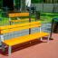 Ryga Bench w/ Backrest, kollased puidust lauad ja hallid metallraamid mänguväljaku ääres.